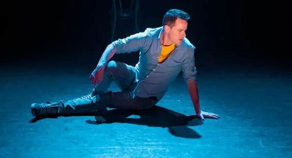A man in denim is sat on the floor of a stage looking away to his left.